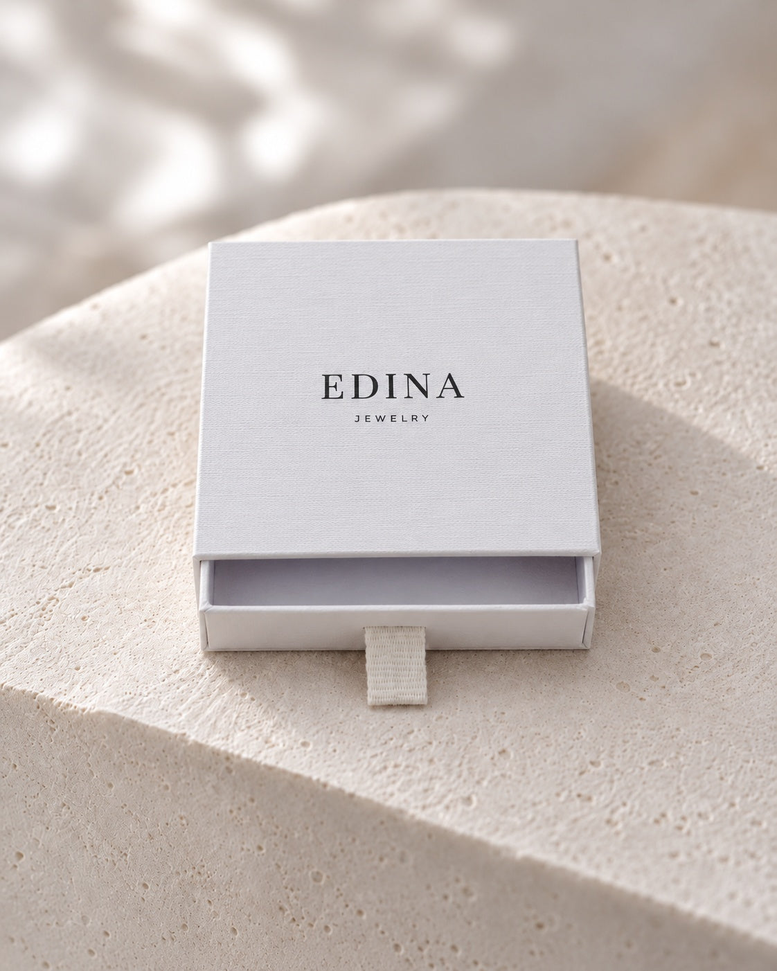 Leicht geöffnete weiße Schmuckbox von Edina Jewelry auf hellem Stein.