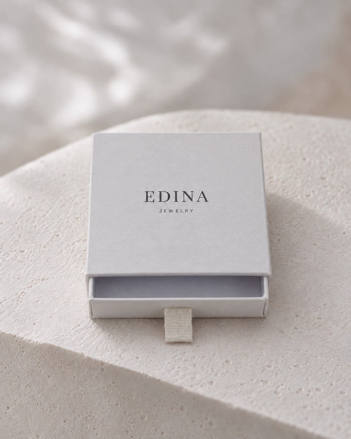 Leicht geöffnete hellgraue Schmuckbox von Edina Jewelry auf hellem Stein.